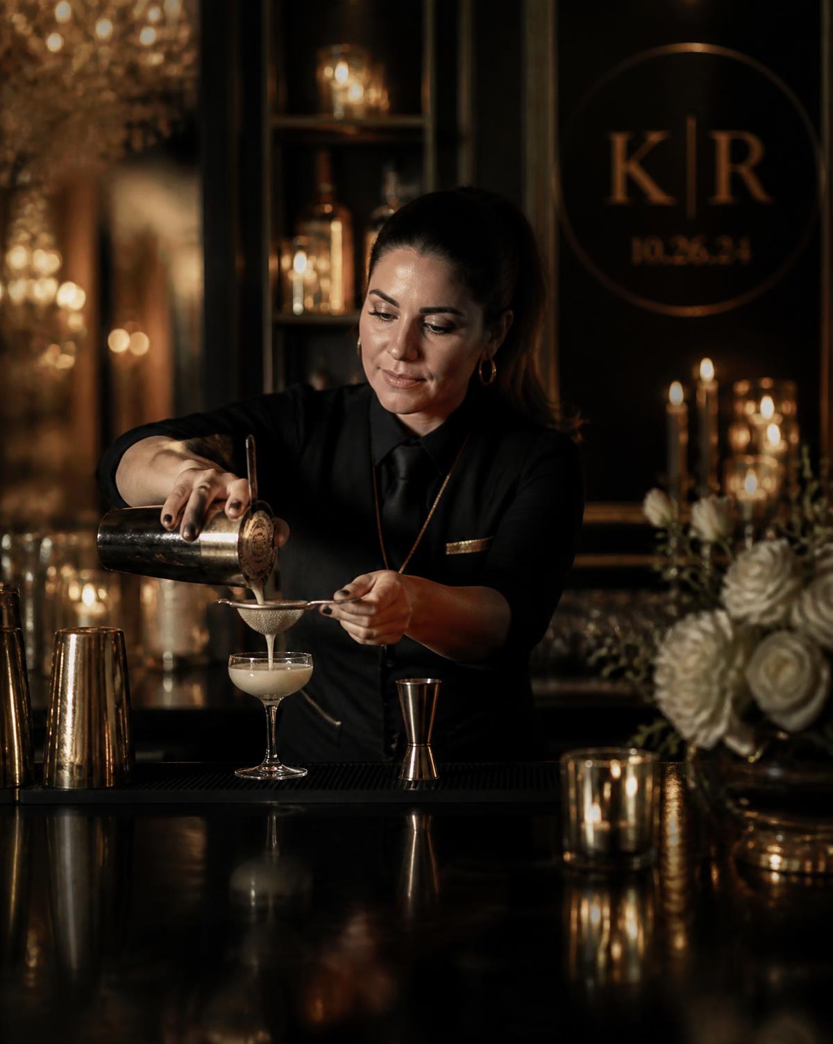 Woman bartender in a black uniform straining a cocktail into a coupe glass